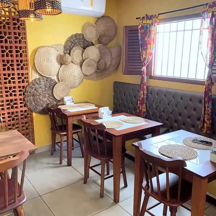Cantinho com mesas de madeira, balaios de palha na parede amarela e banco estofado